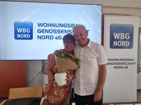 Ein lächelndes Paar steht vor einem großen Bildschirm mit dem Schriftzug WBG NORD.