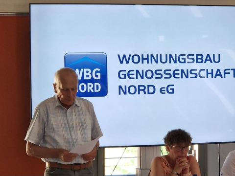 Ein Mann hält eine Präsentation vor einem großen Bildschirm, der das Logo und den Namen einer Wohnungsbaugenossenschaft anzeigt.