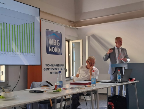 Ein Mann hält eine Präsentation vor einem Balkendiagramm, während eine Frau sitzt und zuhört.